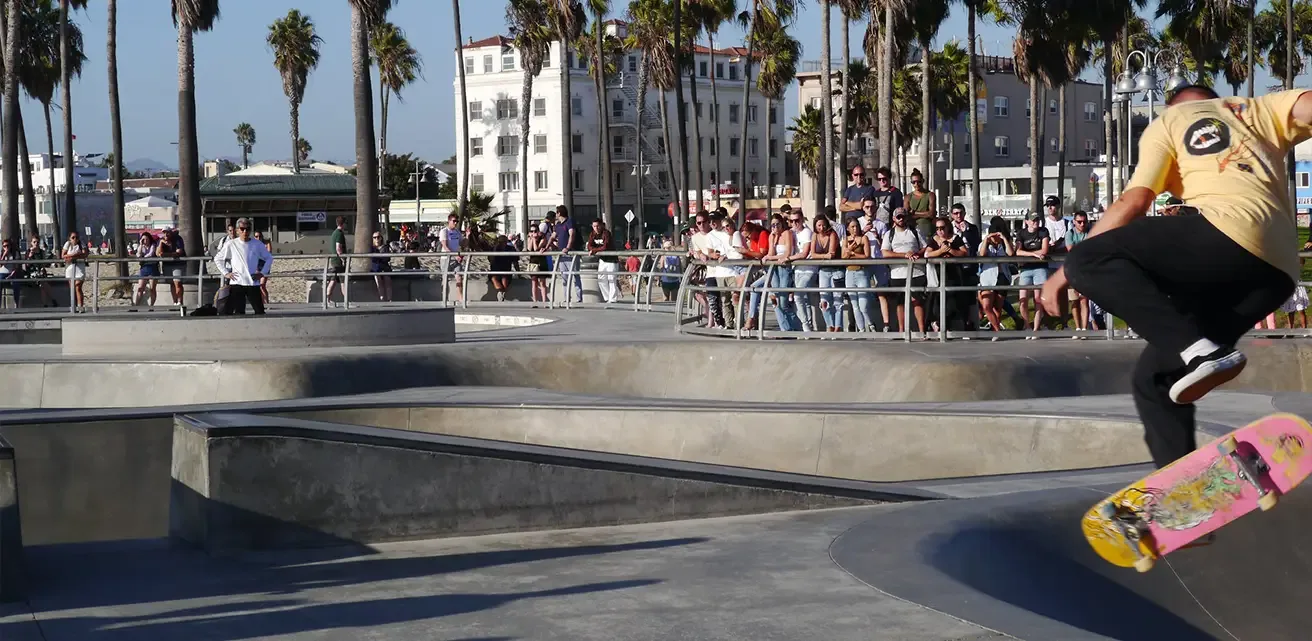 Skateur en plein saut dans un skatepark en bord de plage à Los Angeles, bordé de palmiers et de spectateurs