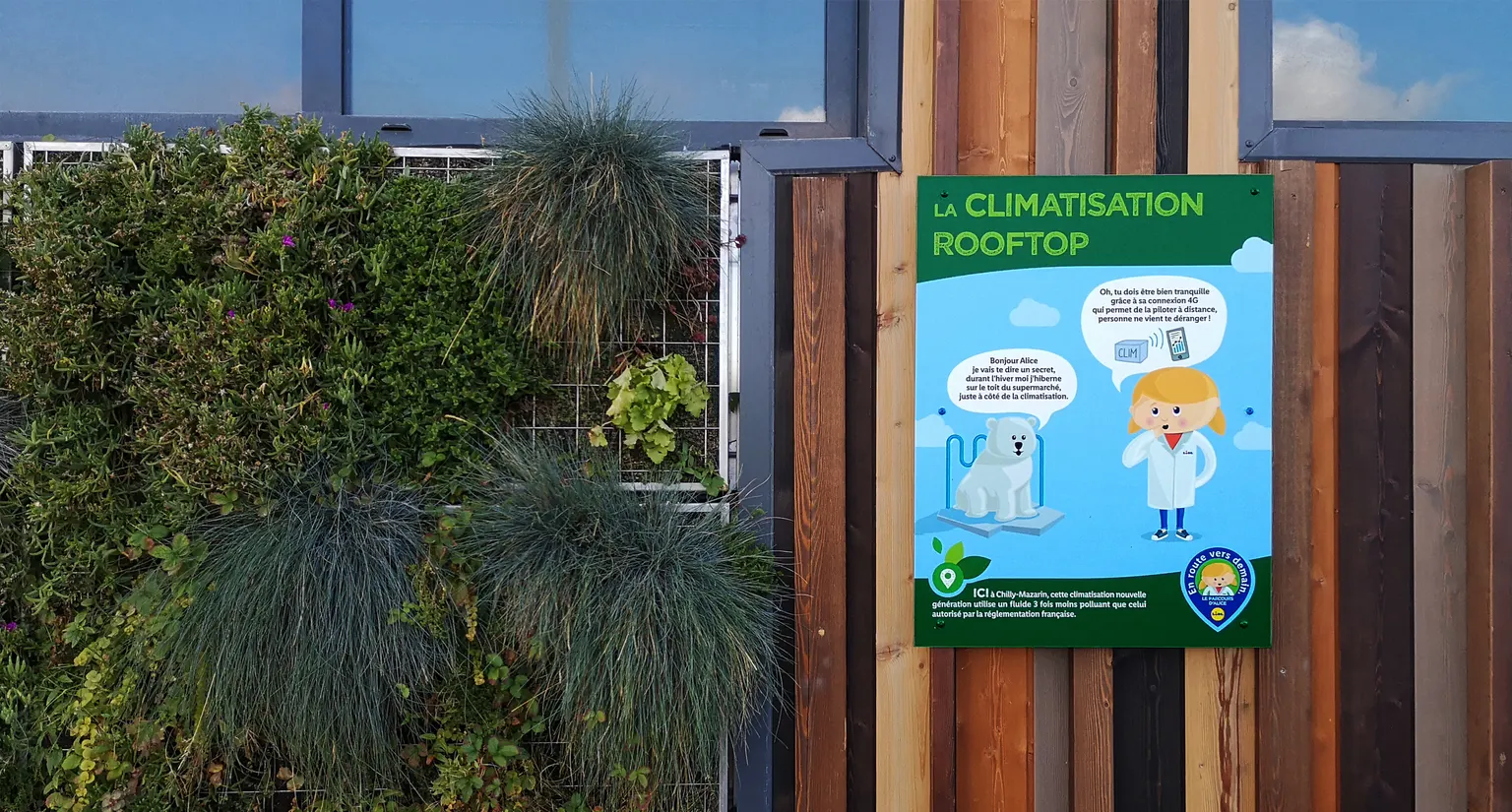 Panneau Lidl La climatisation rooftop illustré du personnage Alice, posé près d'un mur végétal