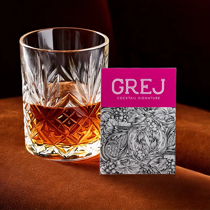 Carte GREJ Cocktail signature rose et gravure de vigne, posée près d'un verre en cristal ciselé